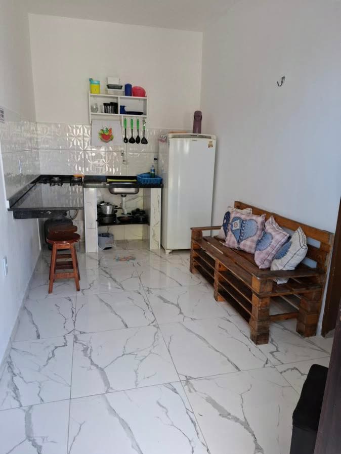 Cozinha de kitnete com geladeira, cooktop sobre bancada preta, prateleira de utensílios e sofá de pallet com almofadas.