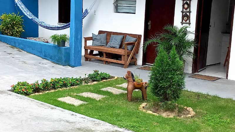 Banco de madeira encostado na parede branca, ao lado de um canteiro de grama com pedras e uma escultura de cachorro.