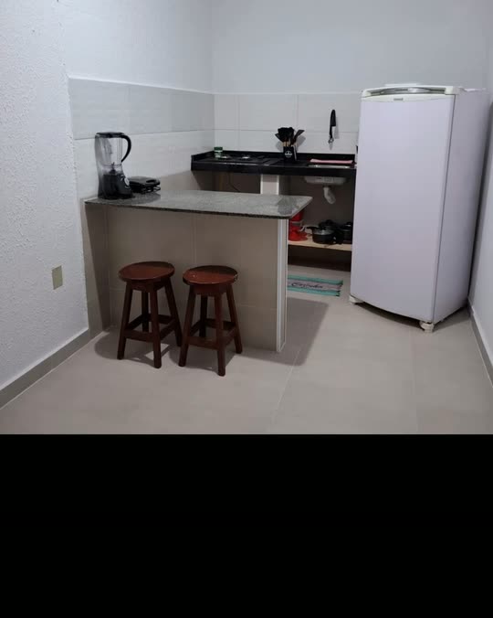 Cozinha compacta da Kitnete Rosa, com cooktop, geladeira branca, bancada e duas banquetas de madeira.