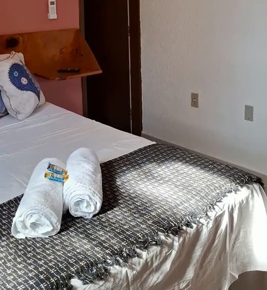 Cama de casal da Kitnete Rosa, com parede rosa, manta cinza e toalhas enroladas sobre a colcha.