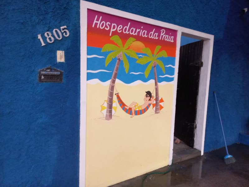 Mural pintado à mão ao lado da porta de entrada, com o nome Hospedaria da Praia, um coqueiro, uma rede e o mar, embaixo do número 1805.