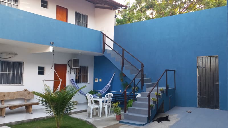 Parede azul da pousada com a escada que leva ao andar de cima, uma rede armada, mesa e cadeiras brancas embaixo.