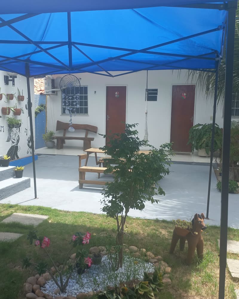 Pátio interno da pousada sob um toldo azul, com coqueiro, canteiro florido, banco de madeira e as portas das acomodações ao fundo.