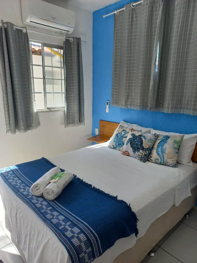 Quarto com cama de casal encostada numa parede azul, roupa de cama branca com faixa azul e janela com cortina clara.