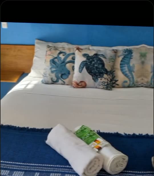 Cabeceira de madeira da Suíte Azul com almofadas estampadas de tartarugas e cavalos-marinhos e toalhas enroladas sobre a cama.