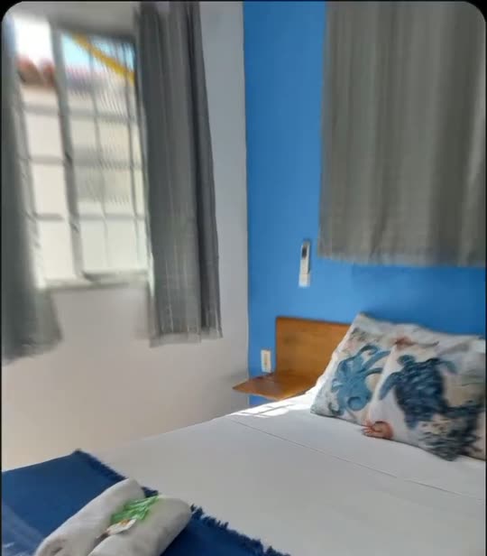 Quarto da Suíte Azul, com cama de casal encostada na parede azul, janela com cortina clara entrando sol e manta azul aos pés da cama.