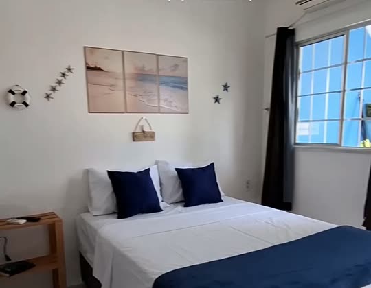 Cama de casal da Suíte Família, com almofadas azul-marinho, quadros de praia na parede e janela ao lado.